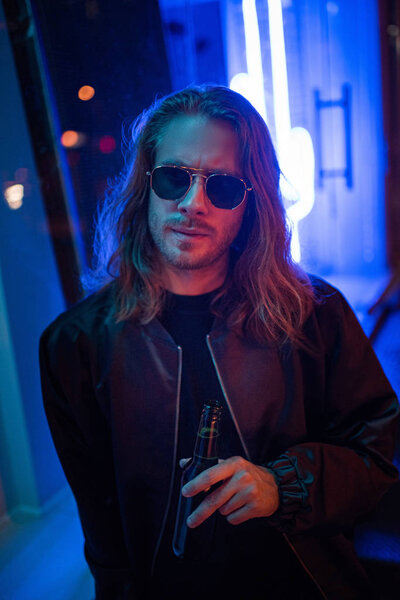 handsome young man in leather jacket and sunglasses with bottle of beer on street at night under blue light