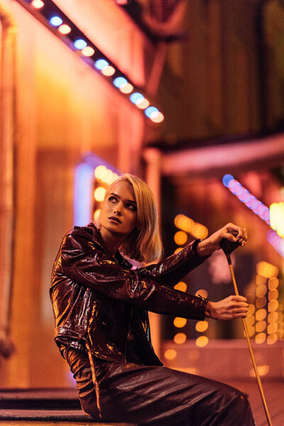 attractive young woman in leather jacket sitting on street at night under yellow light and holding golf club