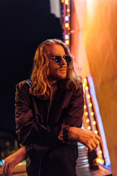 handsome young man in leather jacket and sunglasses on street at night under yellow light