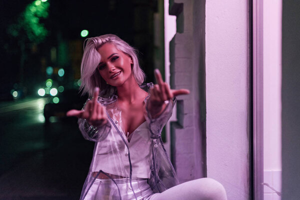 smiling young woman showing middle fingers at camera on street at night under pink light