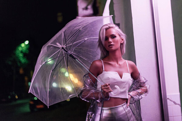 attractive young woman in transparent raincoat with umbrella on street at night under pink light