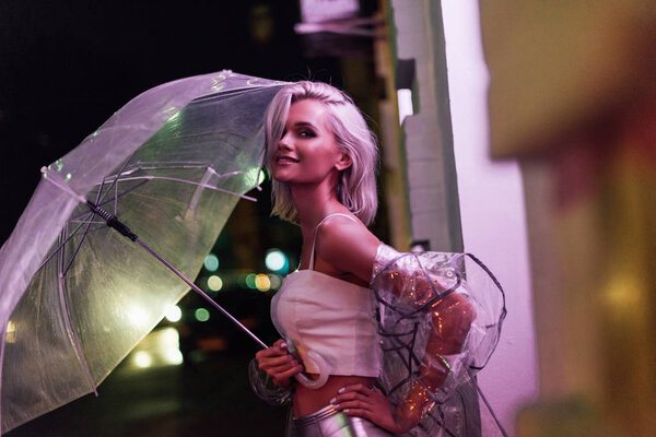 улыбающаяся молодая женщина в прозрачном плаще с зонтиком на улице ночью под розовым светом
