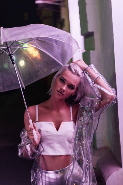 красивая молодая женщина в прозрачном плаще с зонтиком на улице ночью под розовым светом

