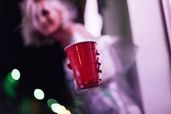 close-up shot of young woman holding red plastic cup on street at night under pink light
