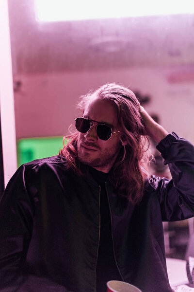 handsome young man in leather jacket and sunglasses spending time on street at night under pink light