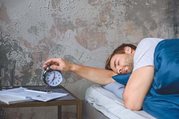 young sleeping man in bed turning off alarm clock at home 