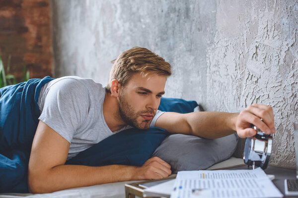 young man waking up in bed and turning off alarm clock at home