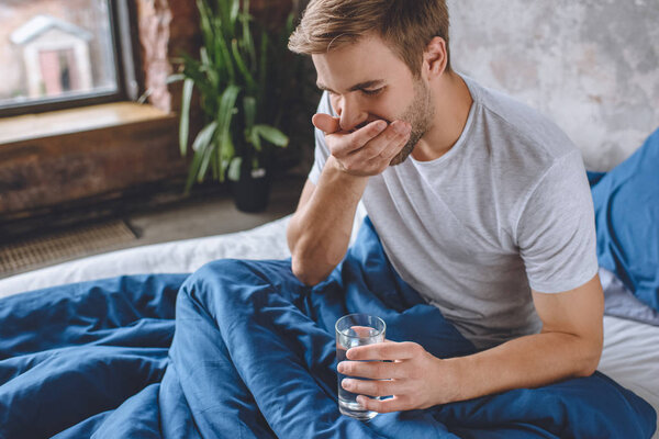 селективное внимание молодого человека, принимающего таблетки и держащего стакан воды в постели дома
