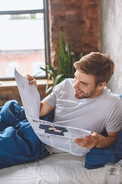 Felice Giovane Uomo Leggendo Giornale Aziendale Letto Durante Mattino Casa — Foto stock gratuita