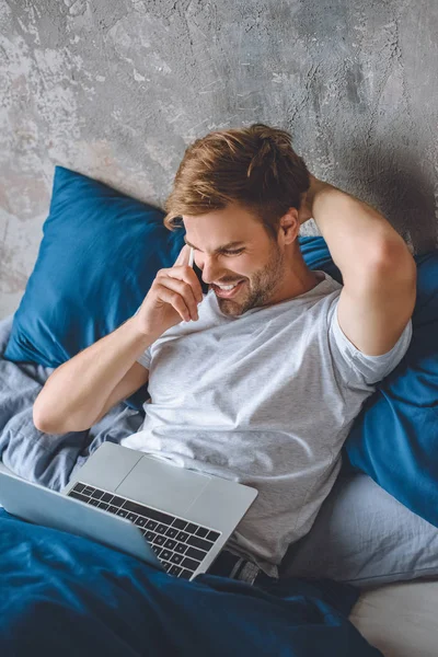 Smiling Young Man Bed Laptop Talking Smartphone Home — Stock Photo, Image