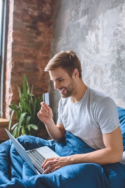 happy young man in bed with credit card doing online shopping on laptop