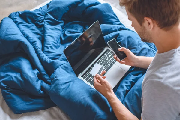 Young Man Bed Credit Card Laptop Doing Online Shopping Smartphone — Stock Photo, Image