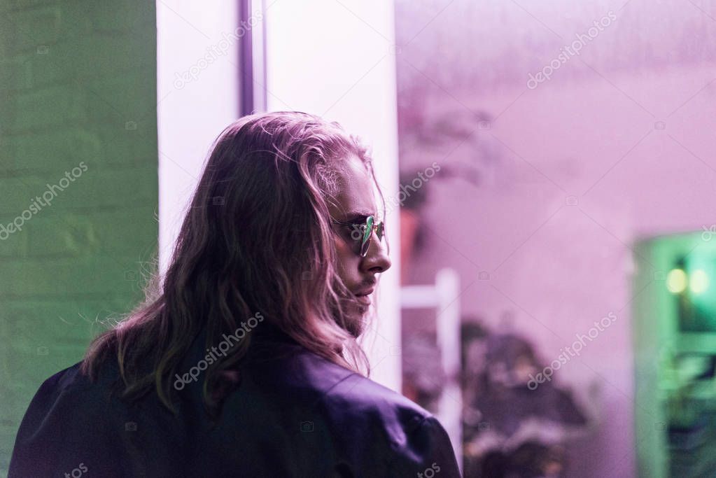 rear view of young man in leather jacket and sunglasses looking away on street at night under pink light