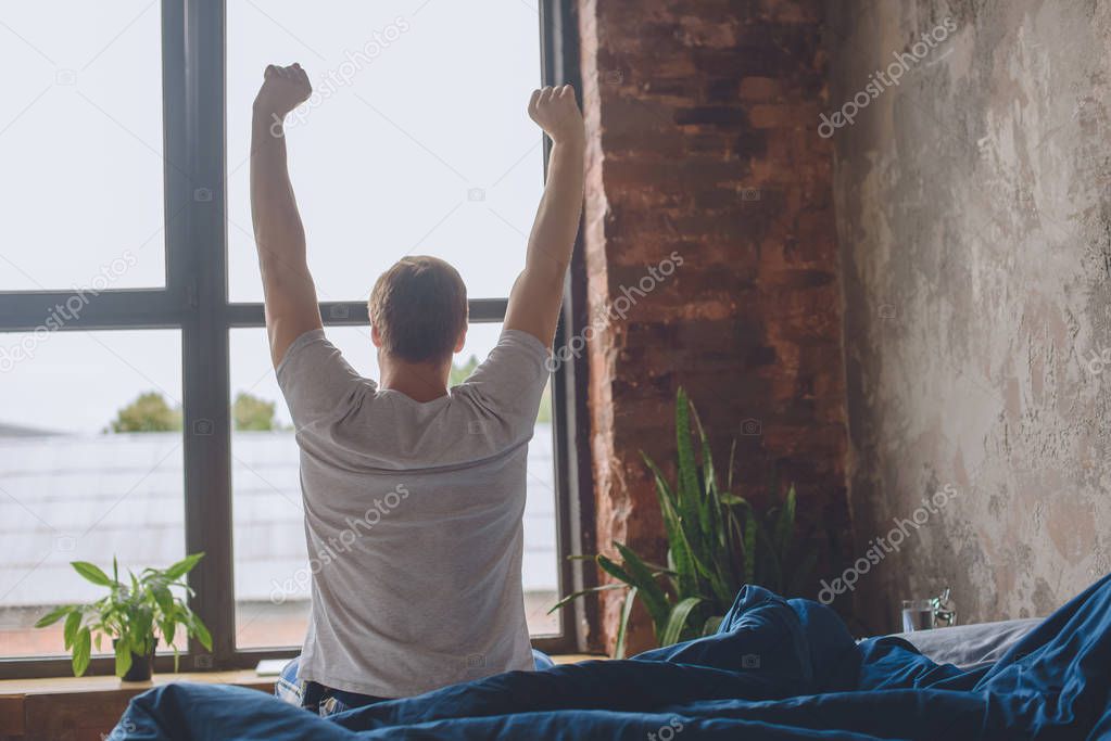 rear view of young man sitting on bed with raised arms during morning time at home 