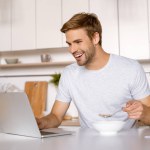 Smiling young man eating flakes with milk and using laptop at kitchen table