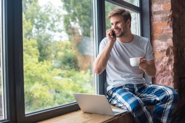 happy young man with coffee cup talking on smartphone while sitting on windowsill with laptop at home clipart