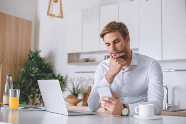 dalgın işadamı Smartphone kullanarak dizüstü bilgisayar ve kahve mutfak masasında kahvaltı sırasında 