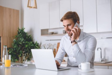 smartphone üzerinde konuşmak ve mutfak masasında dizüstü kullanarak gülümseyen genç işadamı 