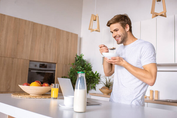 smiling man eating flakes with milk on breakfast and looking at laptop screen at kitchen 