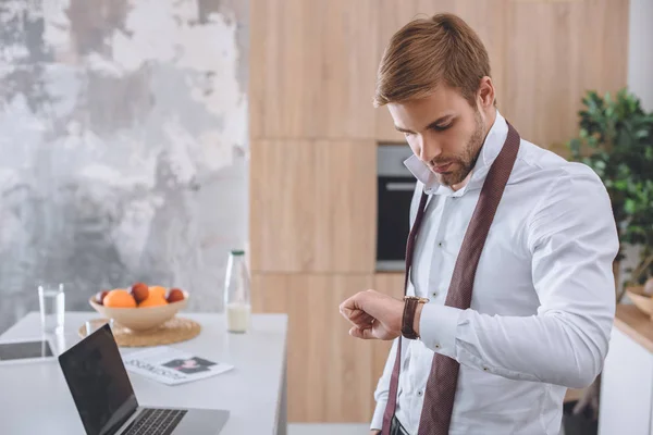 Jungunternehmer Schaut Auf Armbanduhr Küchentisch Mit Laptop — kostenloses Stockfoto