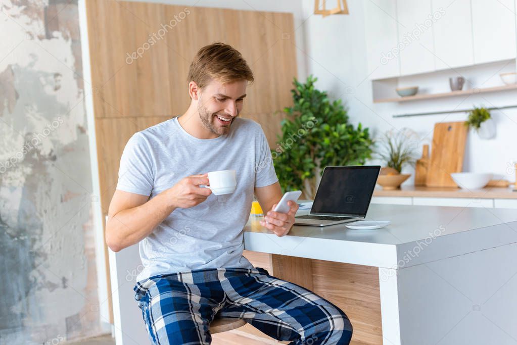 smiling man drinking coffee and using smartphone at kitchen table with laptop