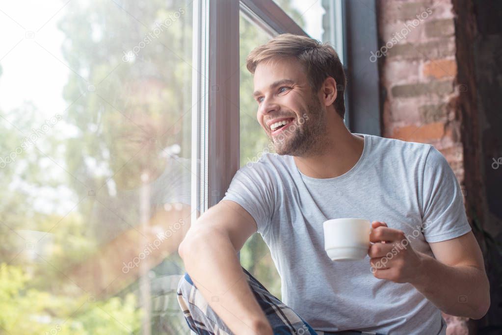 smiling man looking in windows and sitting with cup of coffee on windowsill 