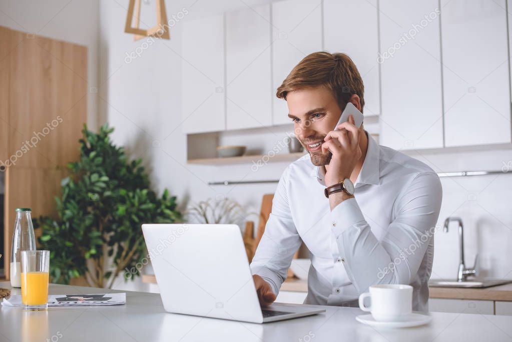 smiling young businessman talking on smartphone and using laptop at kitchen table 