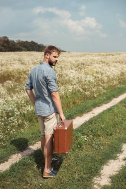 back view of man with retro suitcase standing in filed with wild flowers on summer day clipart