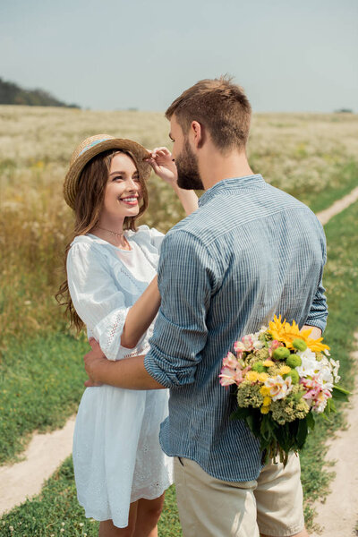 man hiding bouquet of wild flowers for smiling girlfriend behind back in summer field