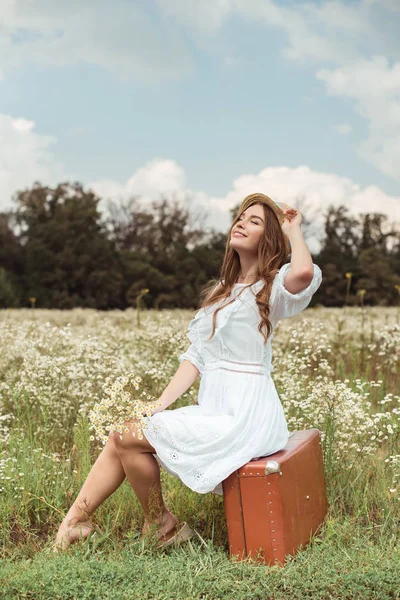 牧草地にレトロなスーツケースの上に座って野生カモミールの花の花束と白いドレスで笑顔の女性 — ストック写真