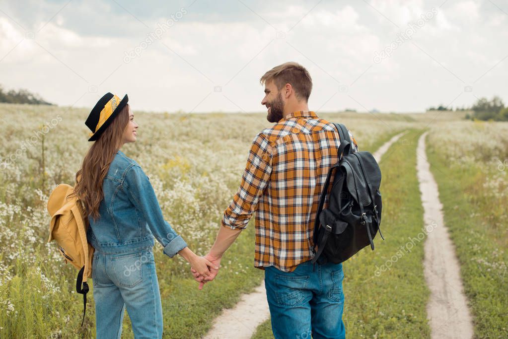 rear view of happy lovers with backpacks holding hands in field with wild flowers