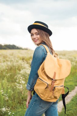 portrait of smiling attractive woman in hat with yellow backpack standing in field clipart