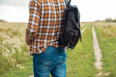 partial view of man in jeans with black leather backpack standing in field clipart