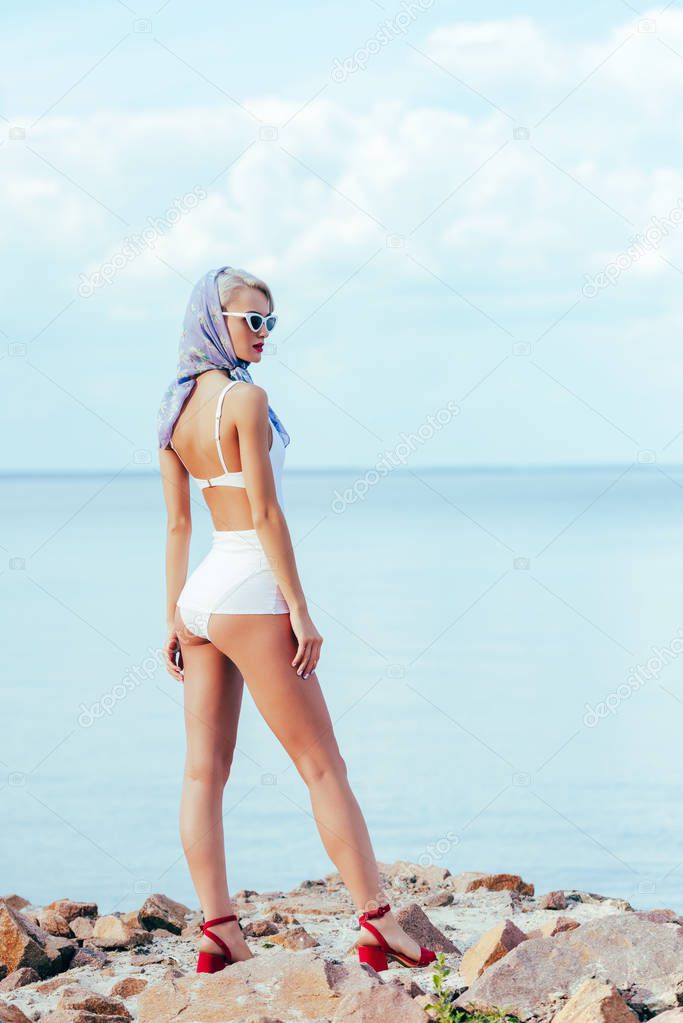 elegant woman posing in white retro swimwear and silk scarf on rocky shore near sea