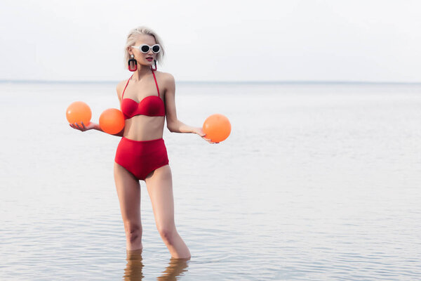 attractive blonde model in red bikini and sunglasses standing in sea and holding balls