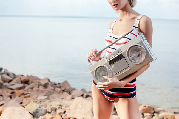 cropped view on girl in striped swimsuit posing with vintage boombox on rocky beach