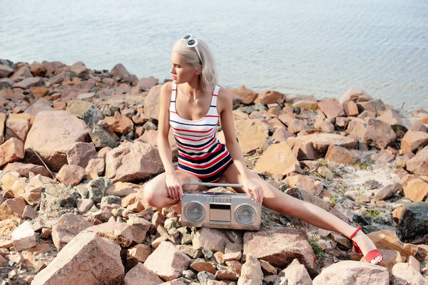 attractive blonde woman in swimsuit sitting on rocky shore with vintage boombox 