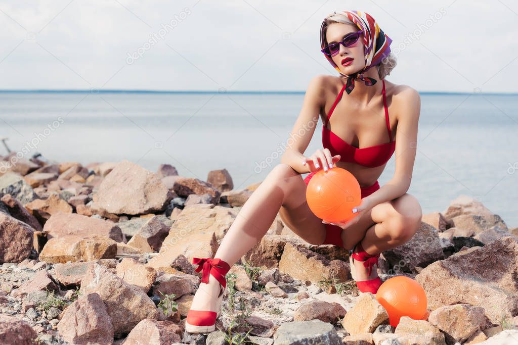 attractive woman in red bikini, sunglasses and silk scarf posing with orange balls on rocky beach