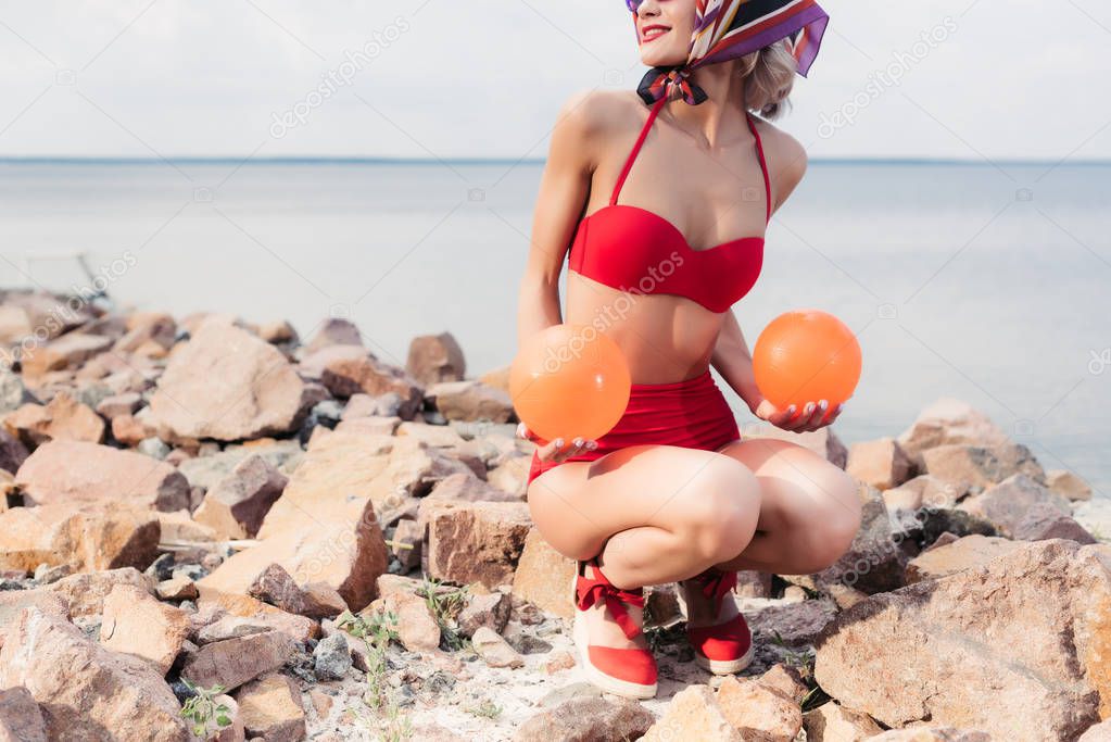 cropped view of woman in vintage red bikini posing with balls on rocky shore