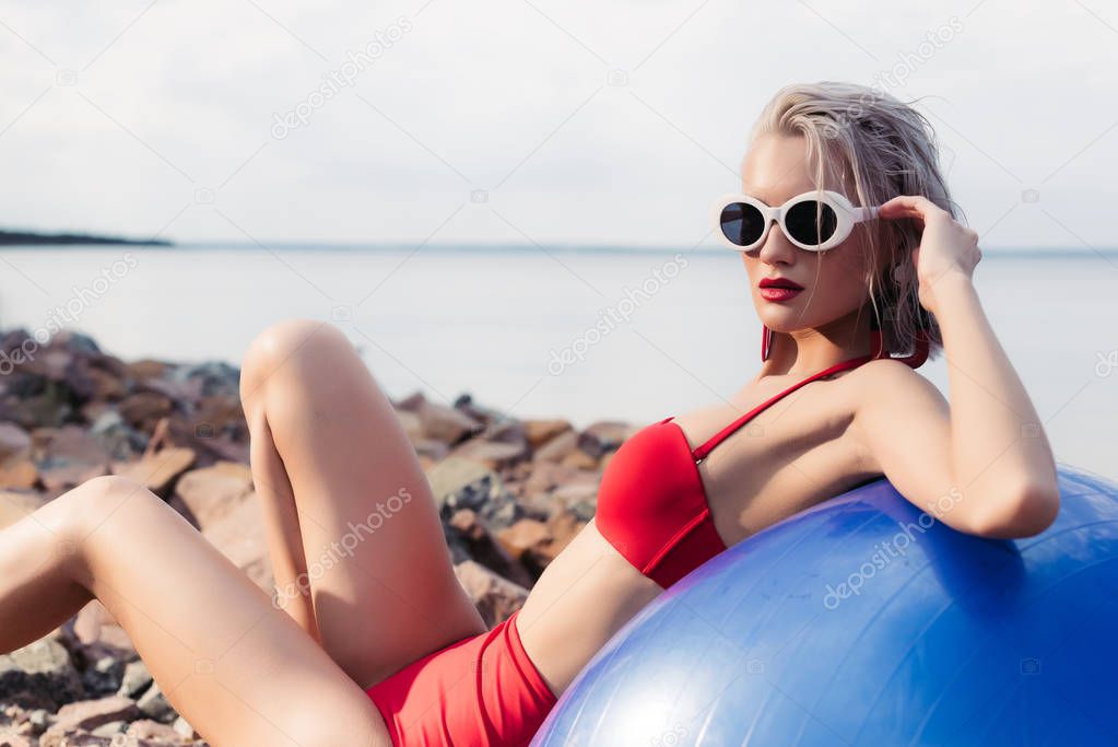 sporty girl in retro red bikini and sunglasses relaxing on blue fitness ball on rocky beach