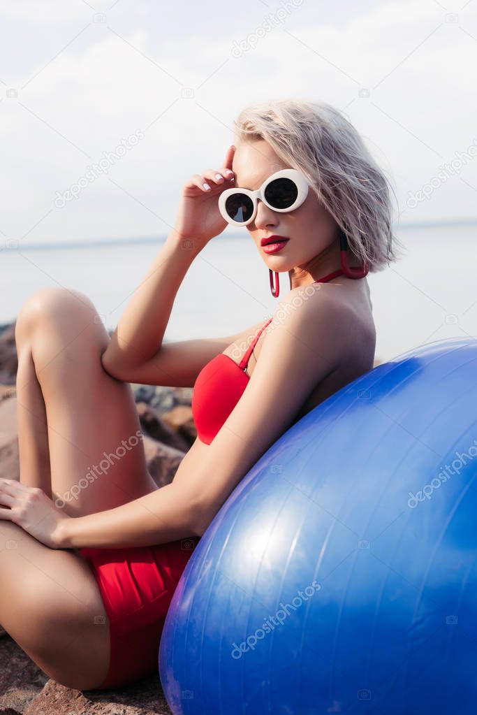 girl in red bikini and sunglasses relaxing on blue fitness ball on rocky beach