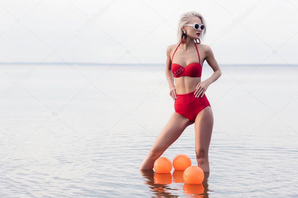 stylish woman in trendy red bikini and sunglasses standing in water with balls