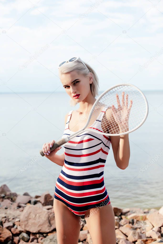 stylish young woman in striped swimsuit posing with white tennis racket on rocky beach