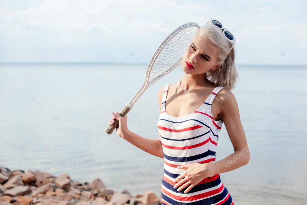 beautiful blonde sportswoman in striped swimsuit posing with tennis racket near the sea