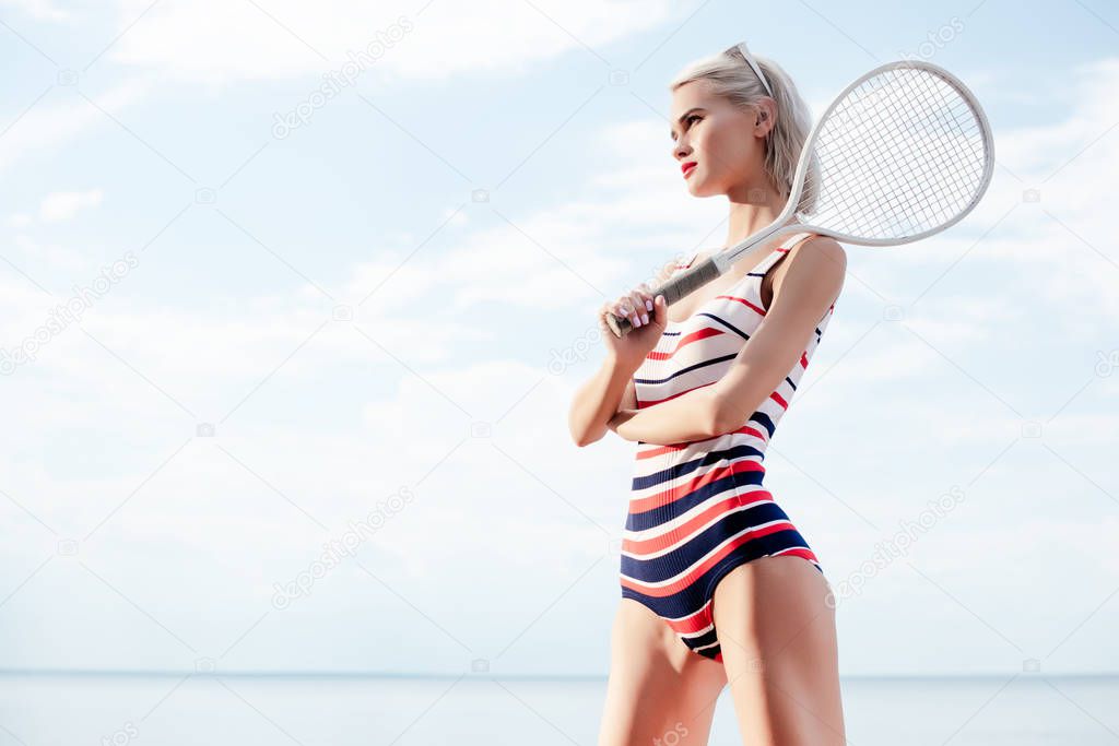 blonde young woman in striped swimsuit posing with white tennis racket near the sea