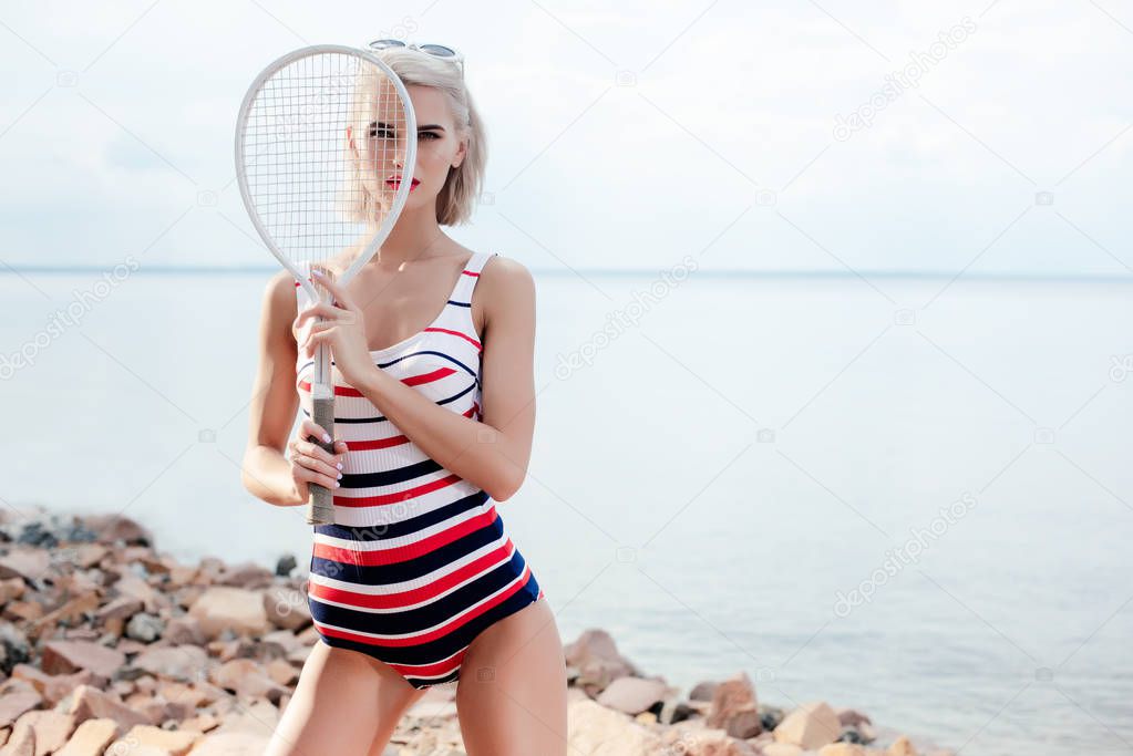 elegant beautiful girl in vintage striped swimsuit posing with white tennis racket on rocky beach at sea