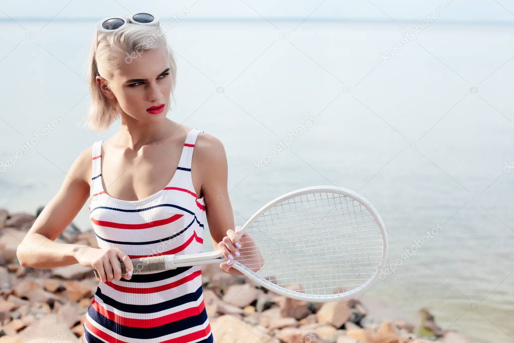 attractive blonde girl in striped swimsuit and sunglasses posing with tennis racket on beach at sea