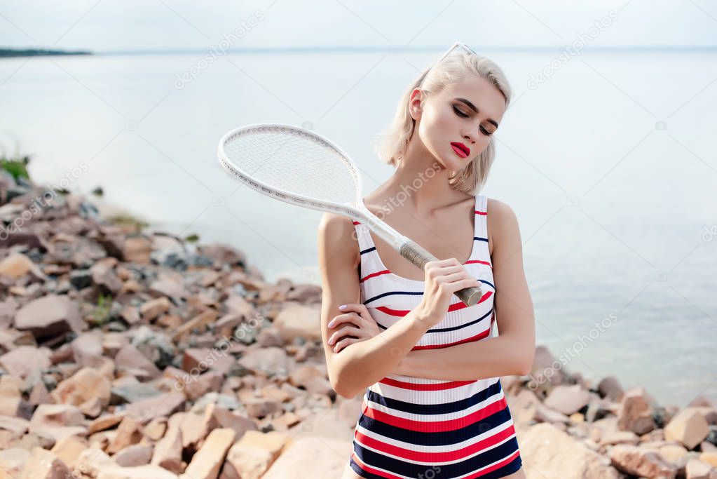 attractive blonde sportswoman in vintage striped swimsuit posing with tennis racket on rocky beach