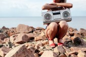 cropped view of stylish woman posing with retro boombox on rocky beach
