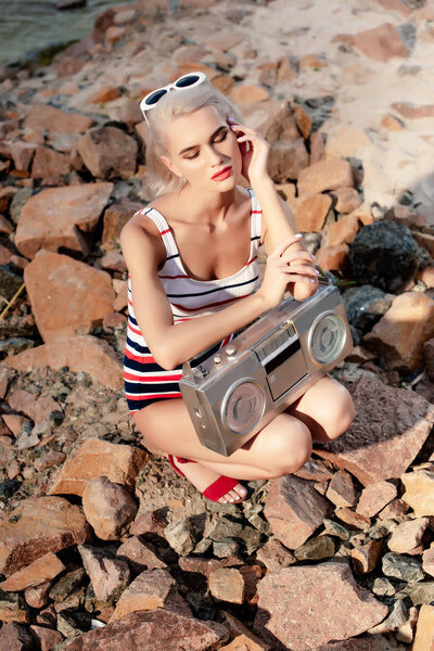 blonde woman in swimsuit posing with vintage cassette recorder on rocks